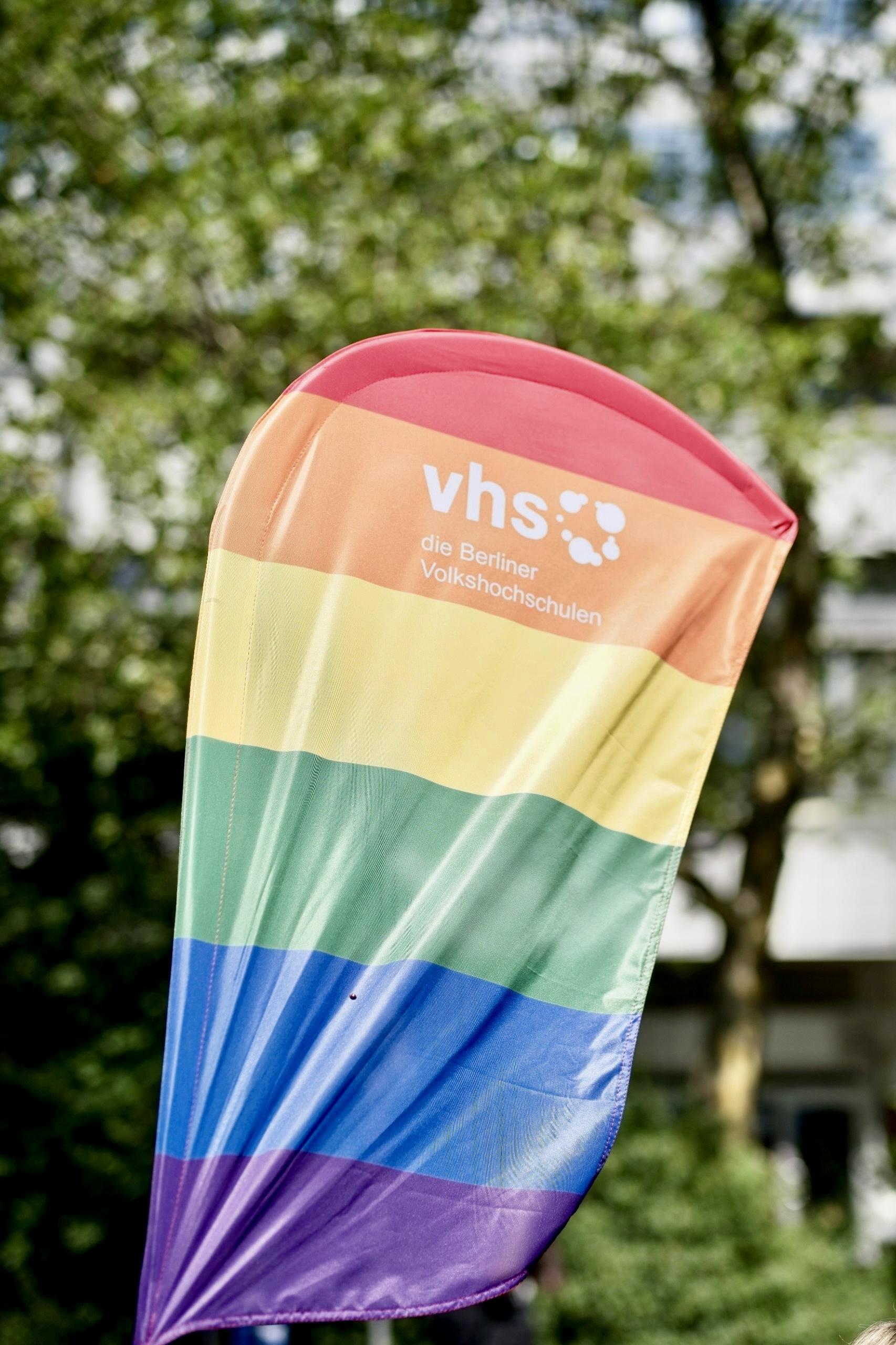 Regenbogenfahne mit weißem vhs Berlin Logo, vor Hintergrund mit Laubbäumen. Aufgenommen beim CSD Berlin, am 25. Juli 2026.