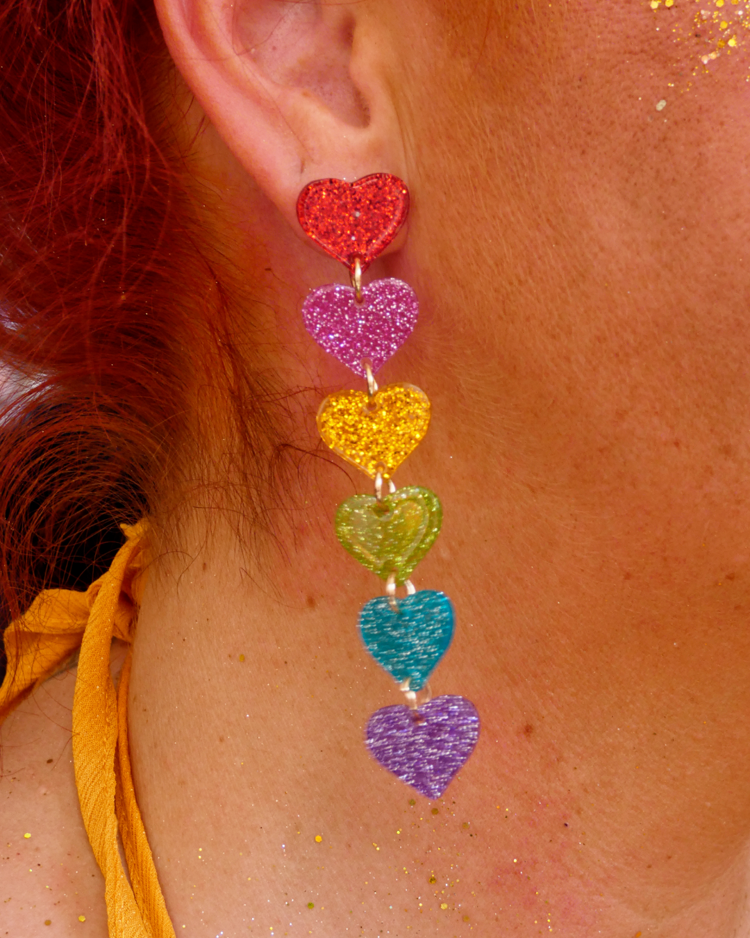 Nahaufnahme eines Ohrrings mit sechs glitzernden Herzanhängern in Regenbogenfarben am Ohr.